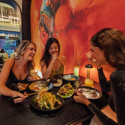 Three women sharing a table of food and laughing, a large red mural on the wall beside them