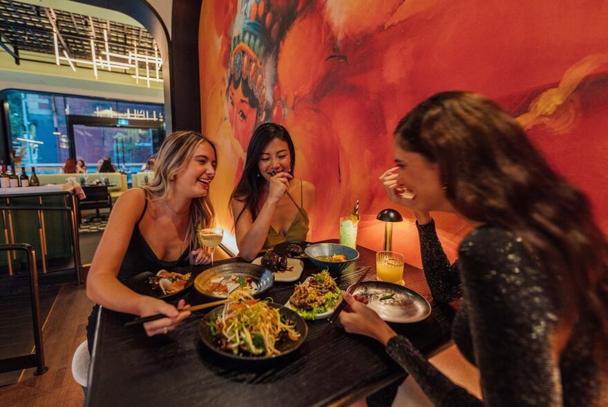 Three women sharing a table of food and laughing, a large red mural on the wall beside them