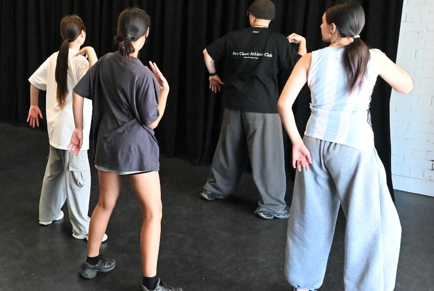 The back of a line of three girls in a dance lesson with a teacher demonstrating at the front. 