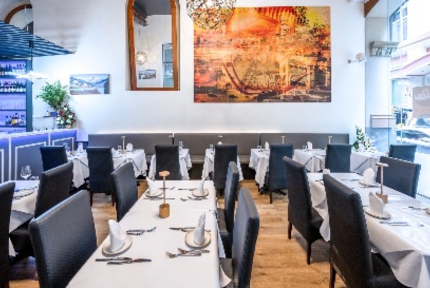 The interior of a restaurant with tables set with white tablecloths and black chairs. A striking red-toned painting on the wall behind.