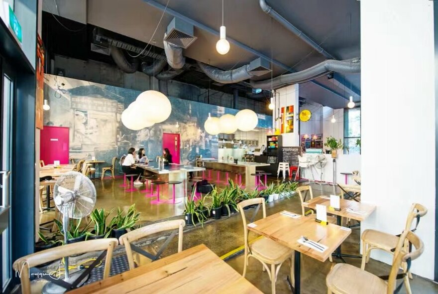 An empty restaurant interior showing wooden tables and chairs, white ball pendant lights and an pedestal fan. 