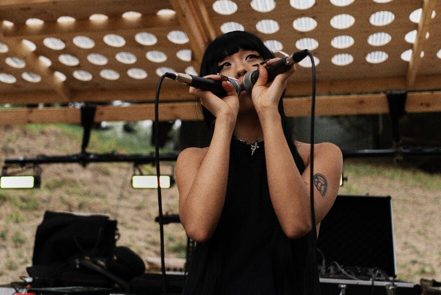 An Asian person wearing black and singing into two microphones on an outdoor stage.