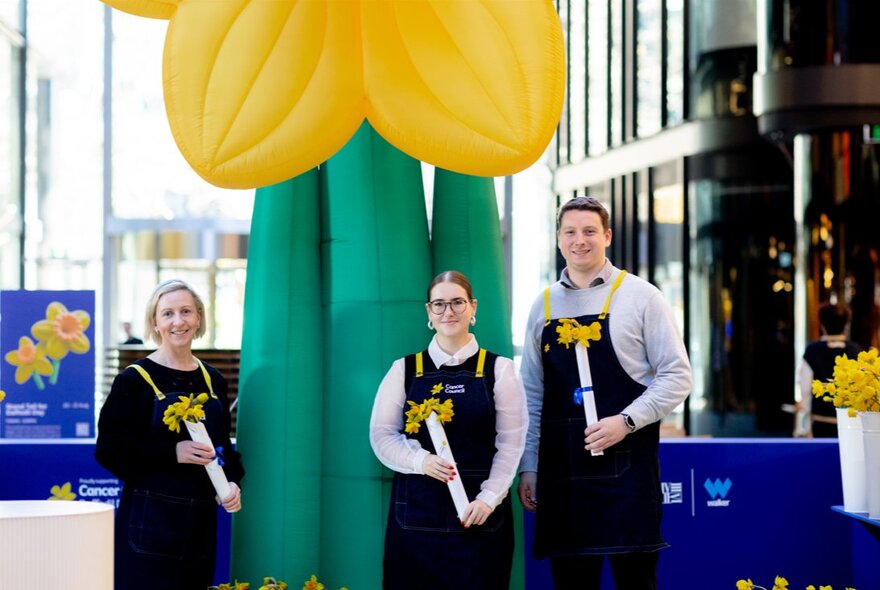 Three people holding daffodils in front of a 4m blow up daffodil.