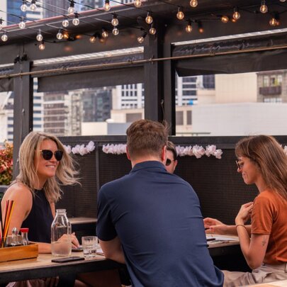 Three people sitting at a table in an outdoor courtyard of a rooftop bar, with city buildings visible in the background.