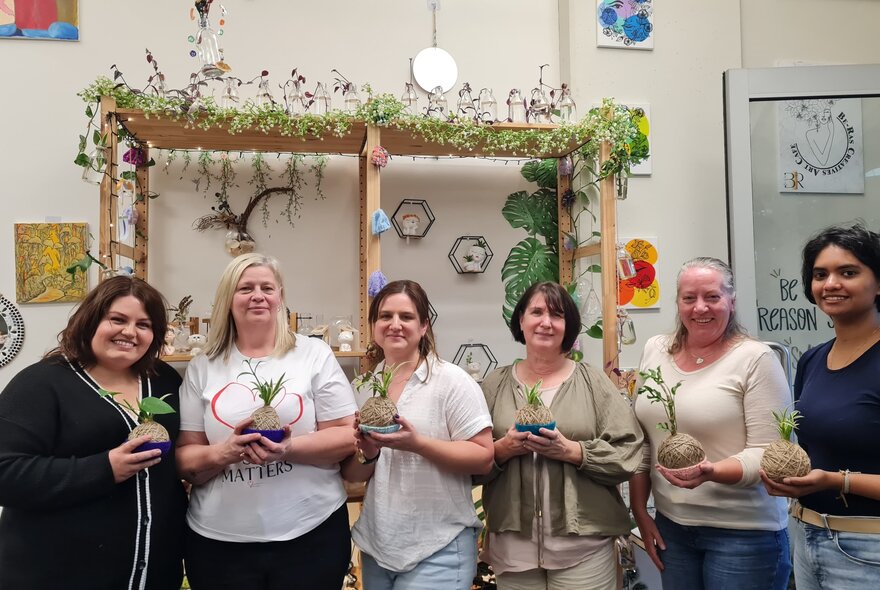 Class participants holding up their home-made kokedama balls of string topped with plants, in a workshop studio room with shelves draped with plants and crafts.