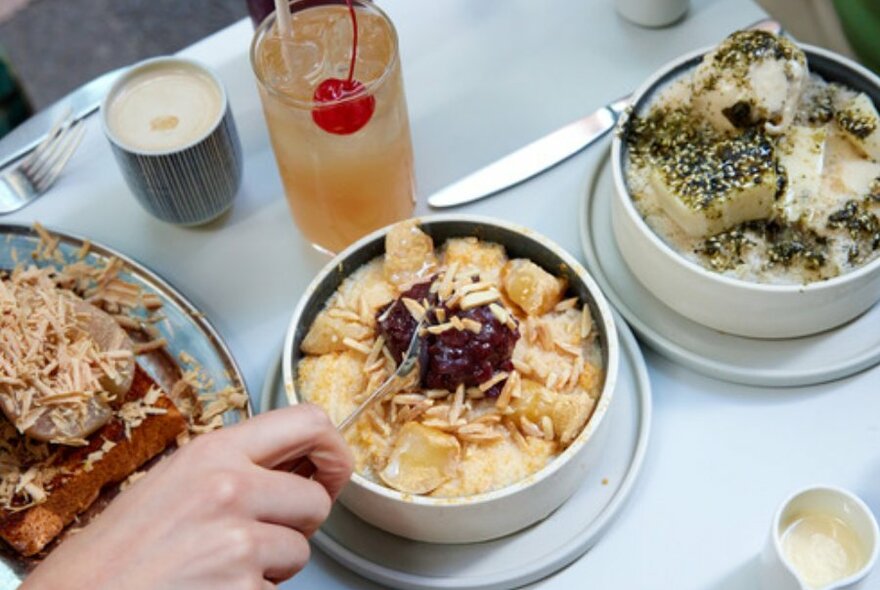 White table with three desserts on it and an iced tea with a red cherry on top. Woman's hand is spooning out yellow dessert with jam.