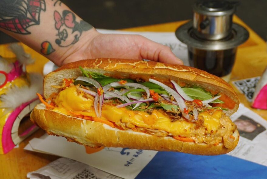 Tattooed hand holding banh mi filled with yellow ingredients and salad.