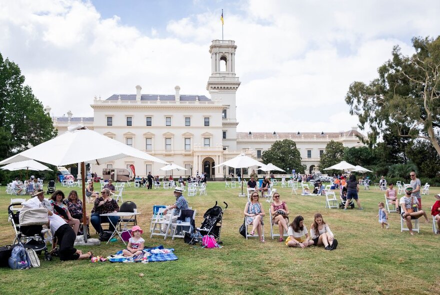 Picnickers seated in deckchairs on the grass under umbrellas in front of the Victorian-era Government House.