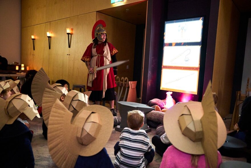 A group of children engaging in an interactive gladiator-themed workshop, the children wearing cardboard helmets, with an adult dressed as a gladiator standing in front of them.