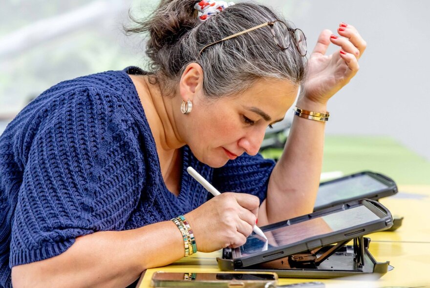A woman leaning over and drawing on an iPad with a stylus, her left hand held upwards near her head. 