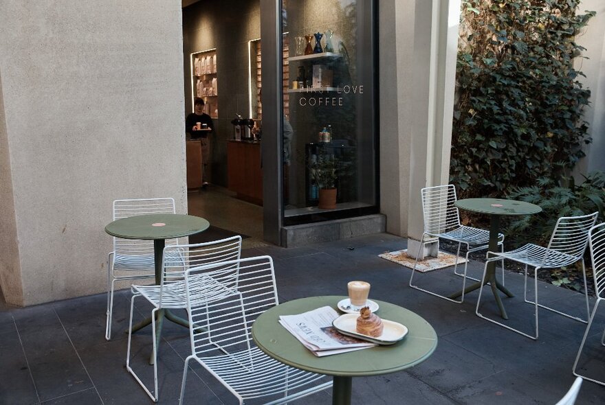 An external courtyard of a cafe with round, sage green tables and white wire chairs; the front table has a coffee, croissant and newspaper on it.