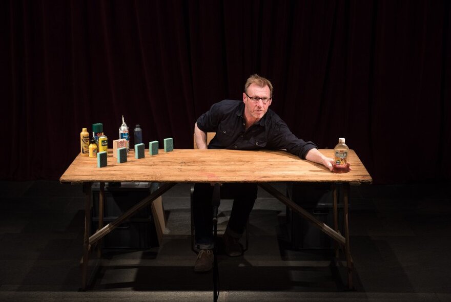A man in black seated at a wooden table on an otherwised darkened stage, reaching out with a sauce bottle, other condiments positioned neatly next to him.