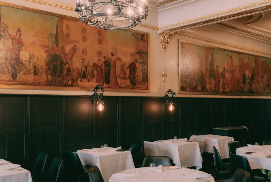 An elegant restaurant dining room featuring dark wood panelled walls beneath a long, historic mural with white tablecloth-covered dining tables neatly arranged across the room.