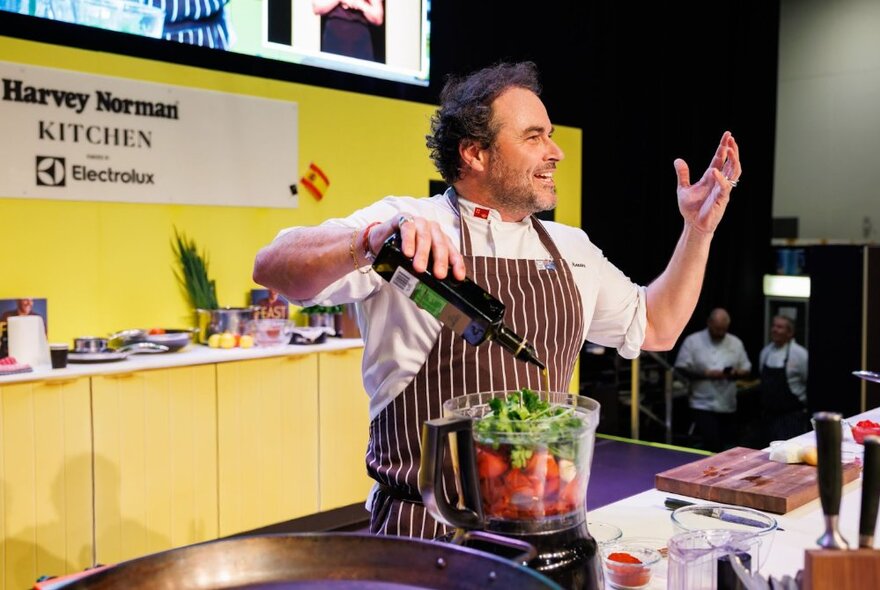 Chef performing live in an event kitchen, smiling and raising his hand while preparing a mixer full of vegetables.