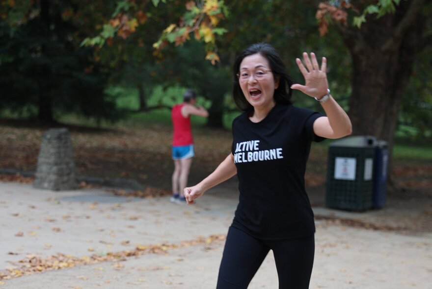 Woman waving in a garden setting, wearing running gear.