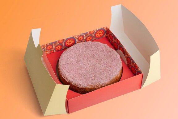 A white gateau cake dusted with pink icing, in a natural red-coloured box with Indigenous designs on the sides.