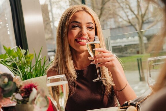 A beautiful young woman enjoying a glass of champagne while seated in front of a window at a restaurant; trees in the background.