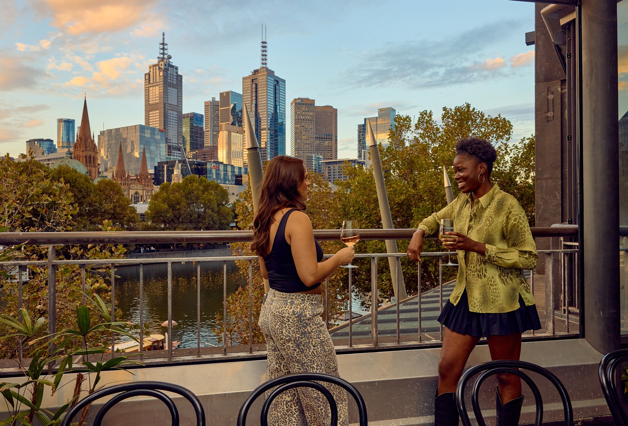 Two friends are drinking wine next to a large open window in a restaurant. There is a view of the Melbourne city skyline and the Yarra River below them.