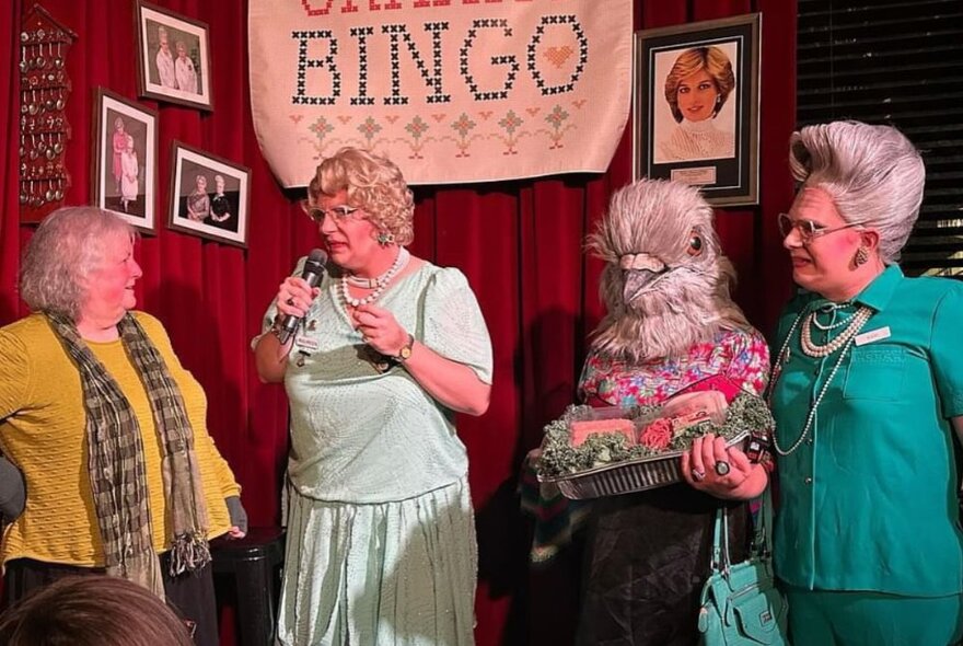 Three dressed as elderly ladies wearing wigs, dresses and jewels interacting with an audience member on stage at Granny Bingo.