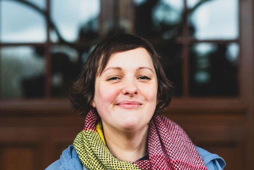 Comedian Josie Long smiling, a scarf wrapped loosely around her neck, standing outdoors in front of a building.