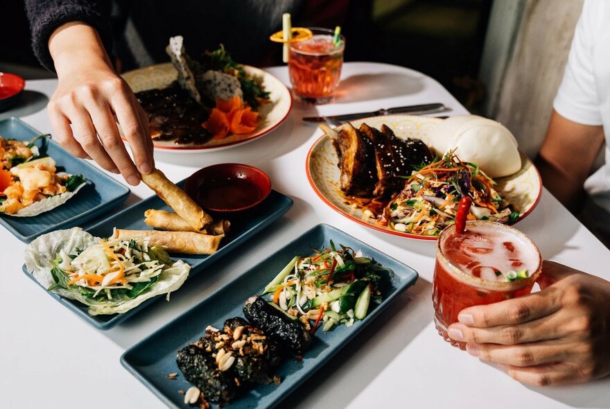 Table laid with several dishes, with hands taking spring rolls from a platter and holding a red drink with ice.