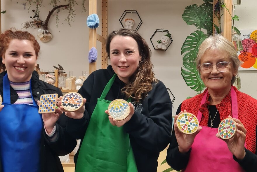 Workshop participants smiling and holding up multicoloured mosaic tiled coasters.