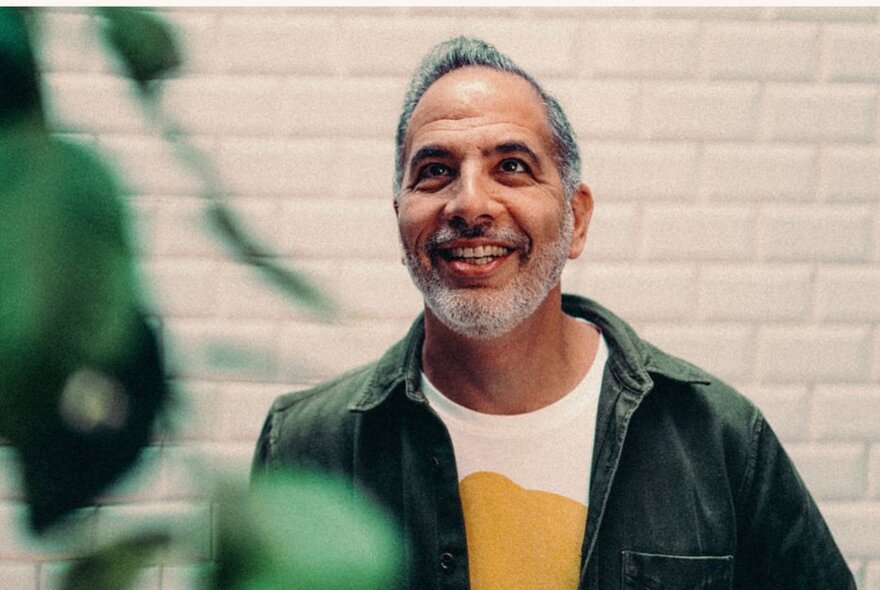 Chef and cook book author Yotam Ottolenghi looking up, wearing a dark green shirt over a white and yellow t-shirt, green plant leaves in the foreground and white brick wall behind him.