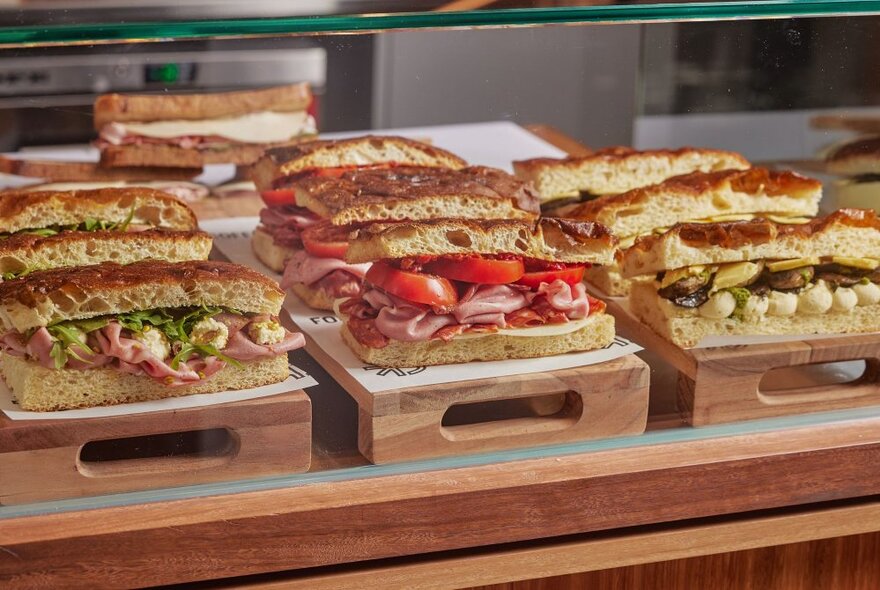 Line-up of large foccacia sandwiches with varied fillings behind a glass and wood counter.