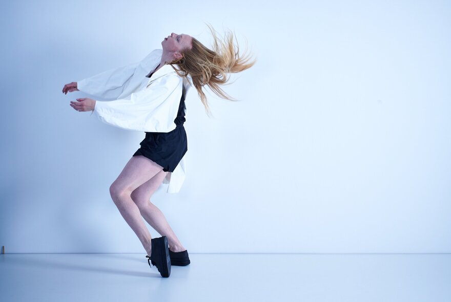 A dancer moving expressively in front of a white wall, her knees bent and her head titled backwards.