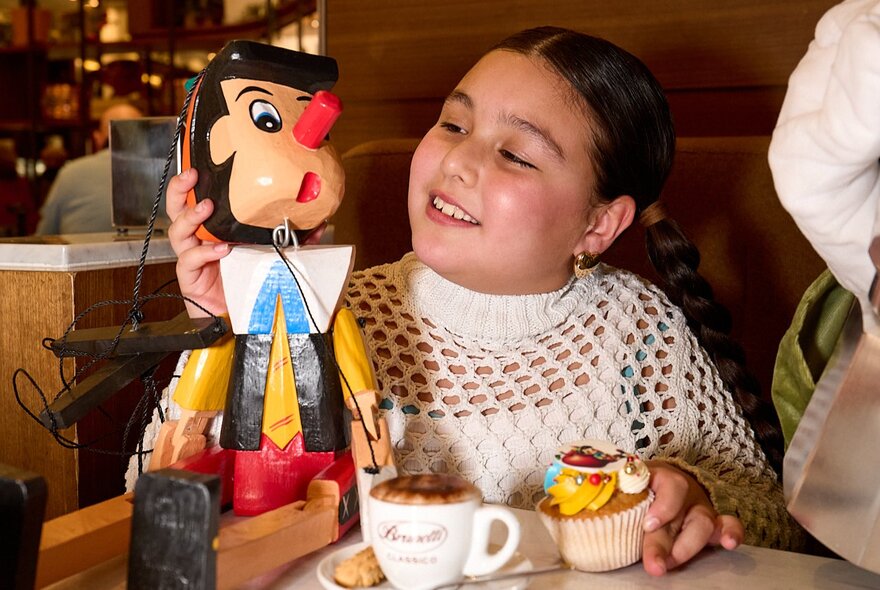A smiling young child seated at a table in Brunetti's Carlton, with cupcakes and a babycino in front of her, she is touching and looking at a Pinocchio wooden doll on the table next to her.