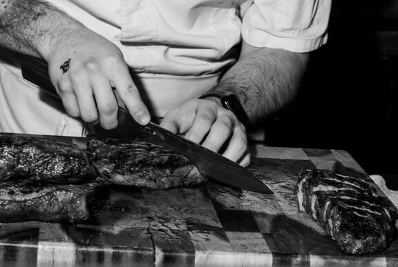 Black and white image of a chef cutting slices from a large steak fillet on a wooden chopping board.