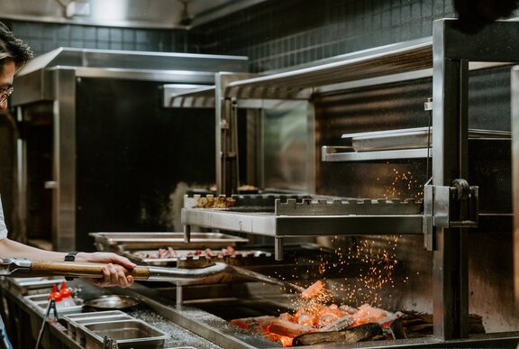 A chef stoking coals with a spade in a commercial kitchen, sparks flying. 