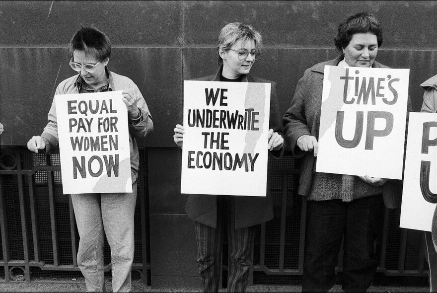 Three women holding up placards with slogans written on them, leaning back against a brick wall on a Melbourne city street; black and white image dating from the mid-1980s.