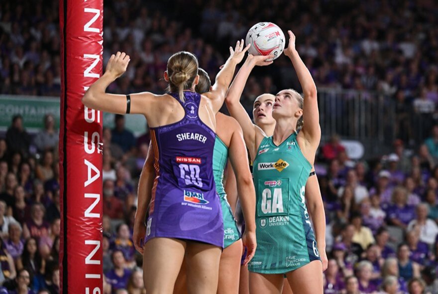 Women's netballers during a game, a player wearing a teal uniform reaching for a goal while a player in purple tries to attack.