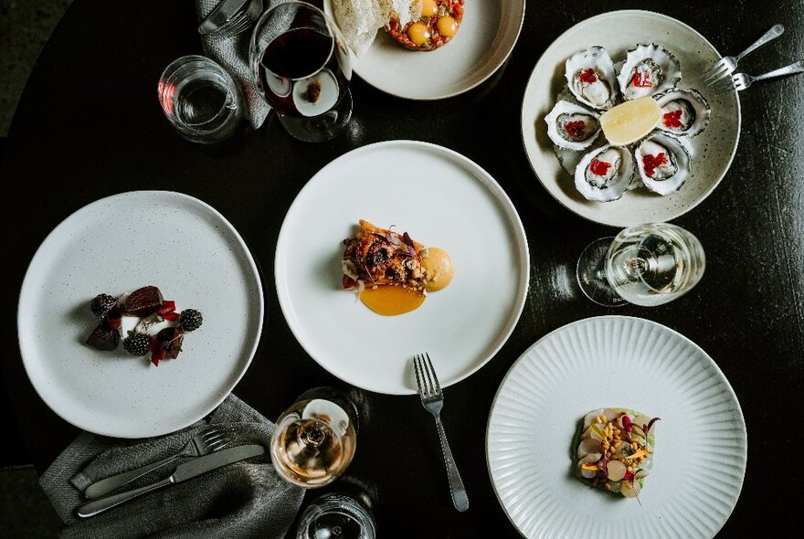 White dishes of gourmet food presented on a black table with cutlery and wine glasses, seen from above.