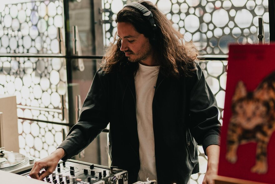 A man with long hair and a mustache wearing a black jacket and white shirt, using DJ equipment in a bar.