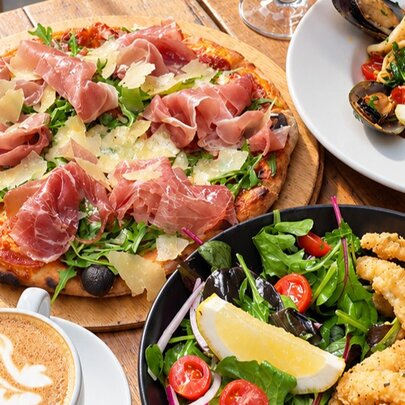 A top-down view of a dining table with a prosciutto and rocket pizza, a seafood pasta dish, a side salad, fried appetizers, and a cup of coffee.