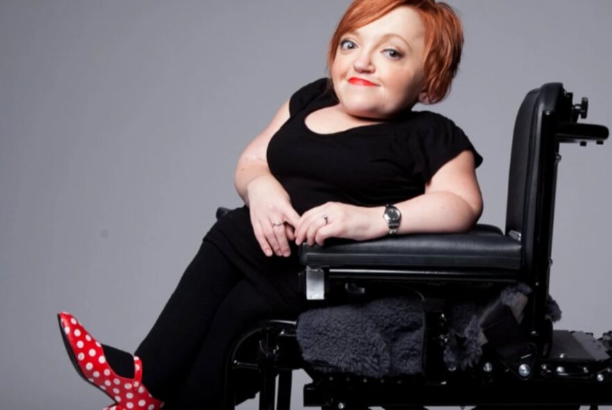 Disabled woman seated in a mobility chair, wearing black, with red polka dot shoes.