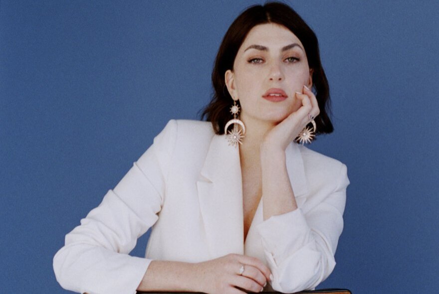 Woman wearing a white suit jacket, her chin is resting in her hand, she has a dark bob hairstyle and is posed against a blue background.