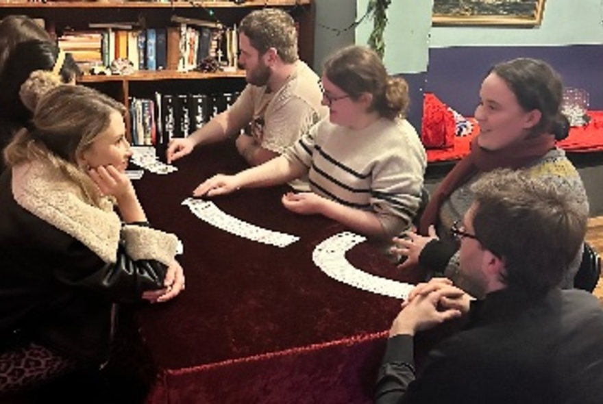 A group of people seated around a dark red velvet covered table, learning how to do card tricks.