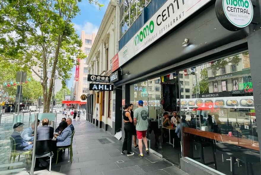 Exterior of Momo Central showing people looking at the menu on display in the window, shop signage and some patrons eating at a table outside on the street.