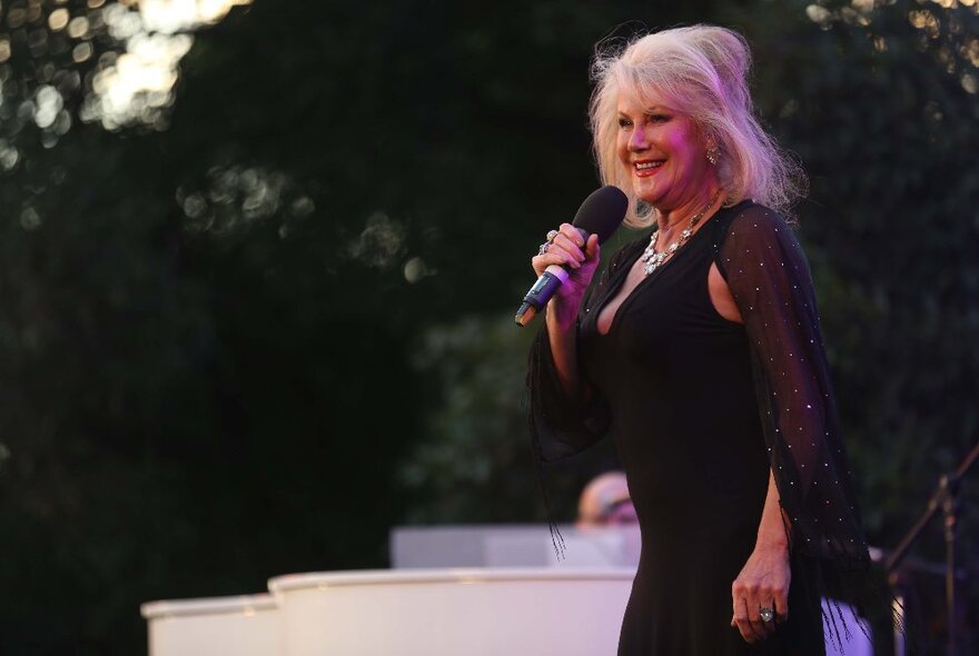 A smiling older woman in a black evening gown, on stage with a microphone outside at dusk.