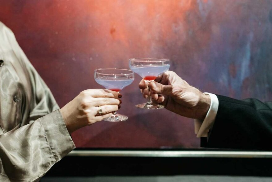 Hands opposite each other bringing two tulip cocktail glasses together in a celebratory clinking motion.