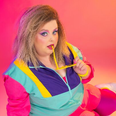 Comedian Mish Wittrup wearing a brightly coloured parachute tracksuit in front of a pinky-orange background.