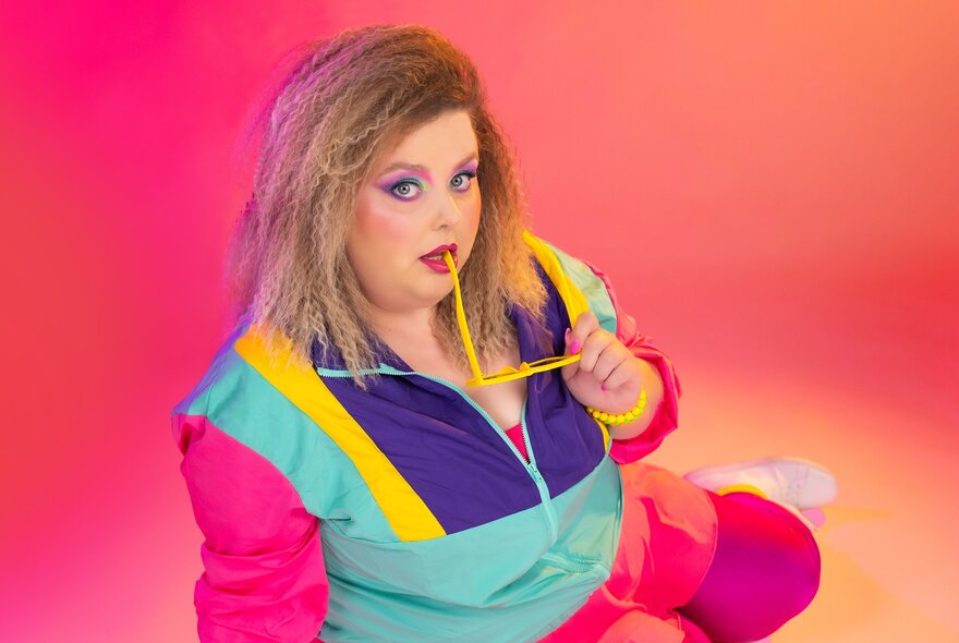 Comedian Mish Wittrup wearing a brightly coloured parachute tracksuit in front of a pinky-orange background.