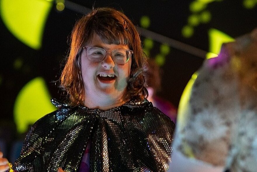 A smiling young woman wearing a glittering cape, with neon lights in the background.