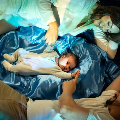A young infant lying on blue satin with its parents lying on either side, touching its hands.