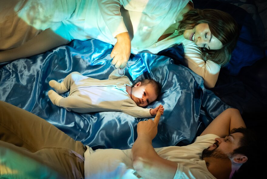 A young infant lying on blue satin with its parents lying on either side, touching its hands.