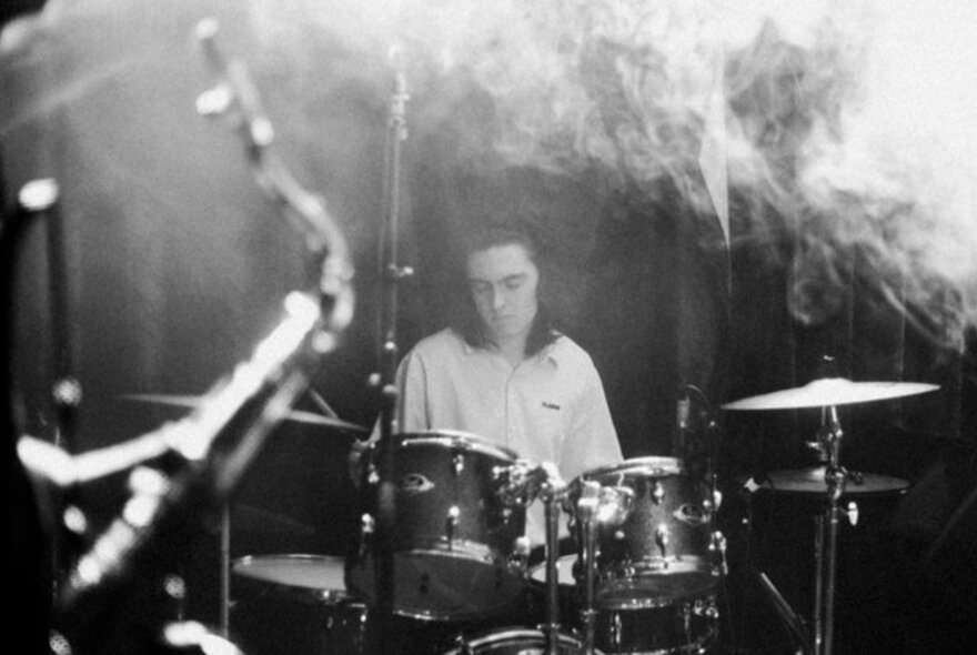 A black and white image of a drummer sitting behind his drumkit, with smoke billowing through the top right corner.