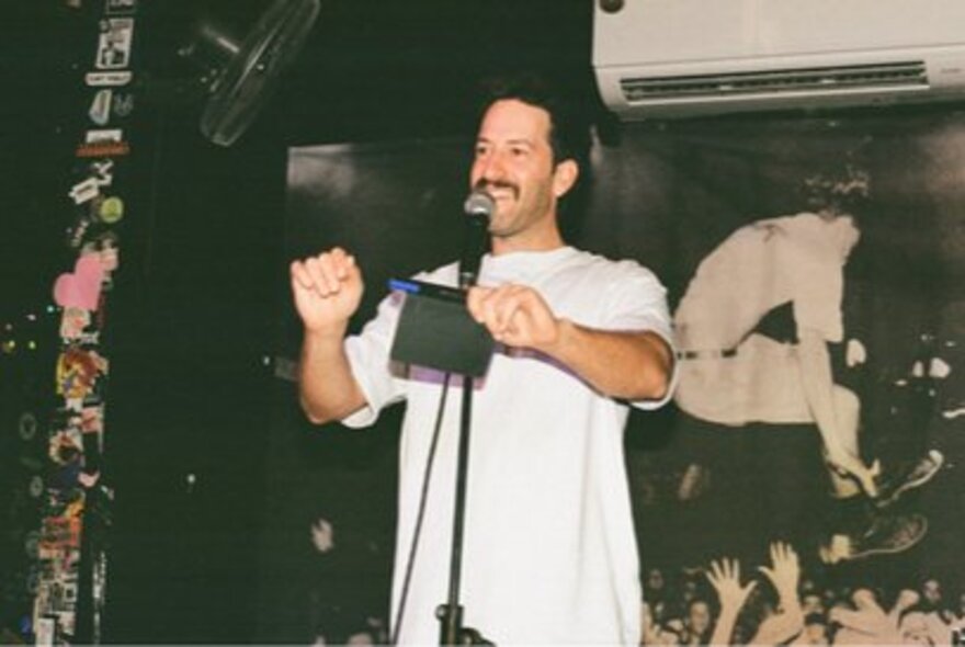 Comedian Ben Kochan on stage performing into a microphone with an split system air conditioner behind him. 